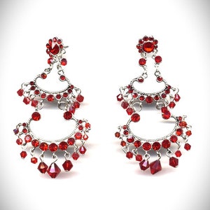May include: A pair of silver dangle earrings with red crystal accents. The earrings feature a tiered design with multiple rows of crystals.