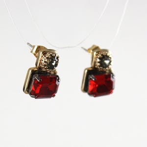 May include: A pair of gold-toned earrings with a square, deep red gemstone at the base. The earrings have a smaller, dark gray stone at the top and are suspended against a white background.