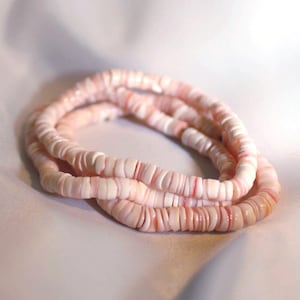 May include: Three strands of pink and white shell bead necklaces. The beads are small, flat, and disc-shaped, strung together to form a delicate, layered accessory. The color palette is soft, with variations of pink and white.