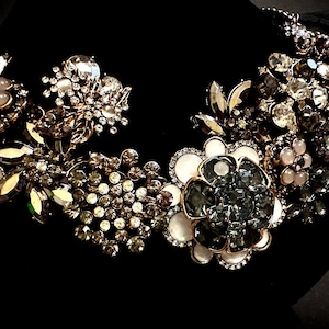 May include: A statement necklace featuring a cluster of floral-shaped components adorned with clear, grey, and pale pink crystals, along with white enamel accents. The design incorporates various metallic settings and faceted stones arranged in a dense, layered pattern.