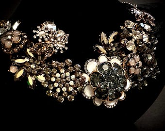 Op de afbeelding: Een statement ketting met een cluster van bloemvormige onderdelen versierd met heldere, grijze en lichtroze kristallen, samen met witte emaille accenten. Het ontwerp bevat diverse metalen zettingen en gefacetteerde stenen in een dicht, gelaagd patroon.