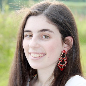 May include: A young woman with long brown hair wearing red and gold dangle earrings. She is smiling and looking at the camera.
