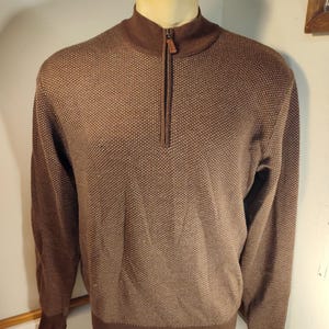 May include: A brown sweater with a textured pattern and a quarter-zip closure. The sweater has a mock neck and long sleeves. The cuffs and hem are a darker shade of brown. The zipper pull is a light brown color.