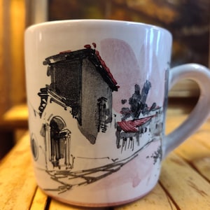 May include: White ceramic mug with a hand-painted illustration of a European village scene. The design features buildings with red roofs, black and gray details, and a pink watercolor background. The mug has a curved handle.