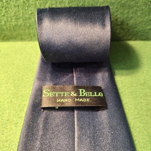 May include: A rolled navy blue tie with a satin finish. The label reads "Sette & Bello HAND MADE" in green lettering on a black background. The tie is displayed on a green surface.