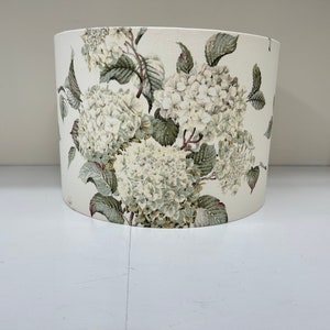 May include: A cream-coloured cylindrical lampshade featuring a floral design. The pattern includes white hydrangea flowers and green leaves, repeated across the surface. The lampshade's shape is a rounded cylinder, suitable for a table or floor lamp.