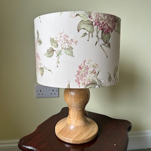 May include: A table lamp with a floral patterned lampshade and a wooden base. The lampshade features a white background with pink and green floral designs. The base is made of light brown wood with a natural wood grain.