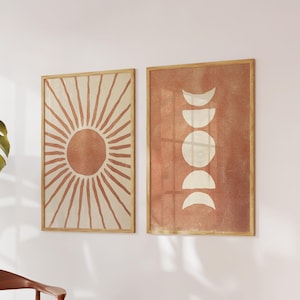 Pode incluir: Duas peças de arte de parede emolduradas. Uma apresenta um design de explosão solar em tons de pêssego e creme. A outra exibe fases da lua em branco sobre um fundo de pêssego. Ambas estão em molduras de madeira clara, criando um conjunto coeso.