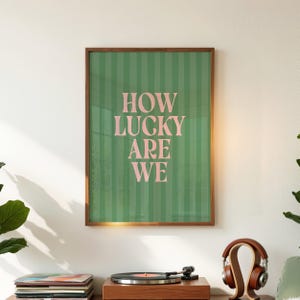 May include: A framed art print with the text "HOW LUCKY ARE WE" in pink letters on a green striped background. The print is in a brown wooden frame. The print is displayed on a wall above a record player and headphones.
