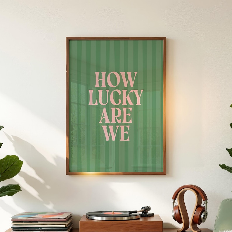 May include: A framed art print with the text "HOW LUCKY ARE WE" in pink letters on a green striped background. The print is in a brown wooden frame. The print is displayed on a wall above a record player and headphones.