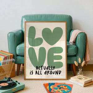 Puede incluir: Una impresión artística enmarcada con la palabra "LOVE" en letras verdes grandes y una forma de corazón, con la frase "ACTUALLY IS ALL AROUND" en texto negro. La impresión se exhibe en una habitación con un sillón verde azulado, un tocadiscos y discos de vinilo.