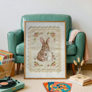 May include: Framed artwork featuring a brown rabbit holding a roll of toilet paper. The artwork is framed in a light wood frame and surrounded by a cream-colored lace border with floral accents. The artwork is displayed in a room with a teal armchair.