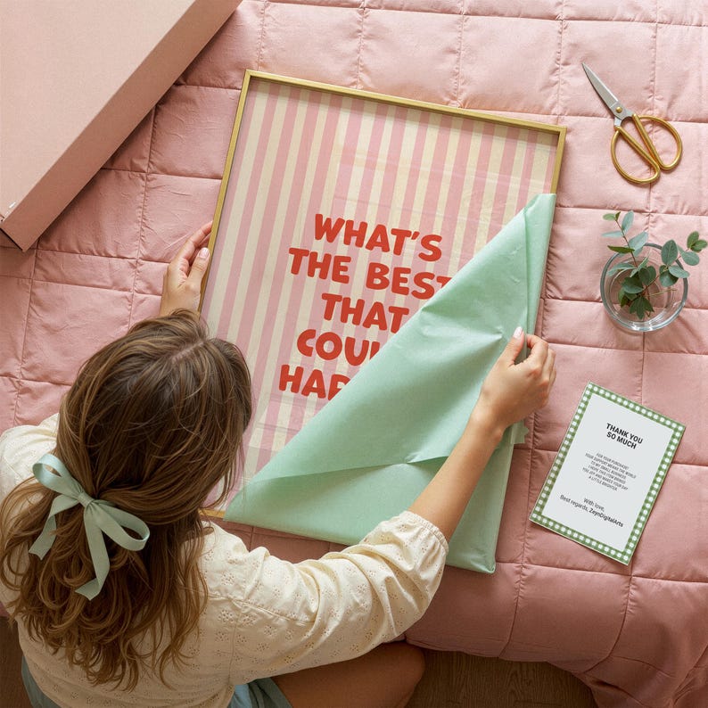 May include: A framed print with a gold border, featuring vertical pink and white stripes and the text "WHAT'S THE BEST THAT COU' HAPP". The print is being unwrapped from mint green tissue paper. A pink box, scissors, and a thank you card are also visible.