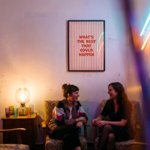 May include: Two people are seated in patterned armchairs, engaged in conversation. A framed poster on the wall reads "WHAT'S THE BEST THAT COULD HAPPEN" in red text on a pink striped background. A lit lamp and colorful glasses sit on a wooden side table.