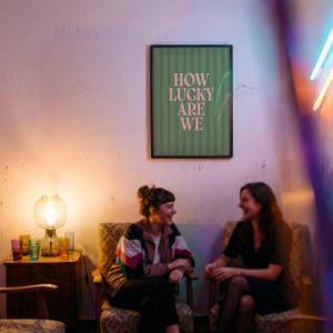 May include: Two people seated in patterned armchairs are laughing. A framed poster on the wall reads "HOW LUCKY ARE WE" in white text on a green striped background. A lit lamp and colorful glasses sit on a small wooden table.