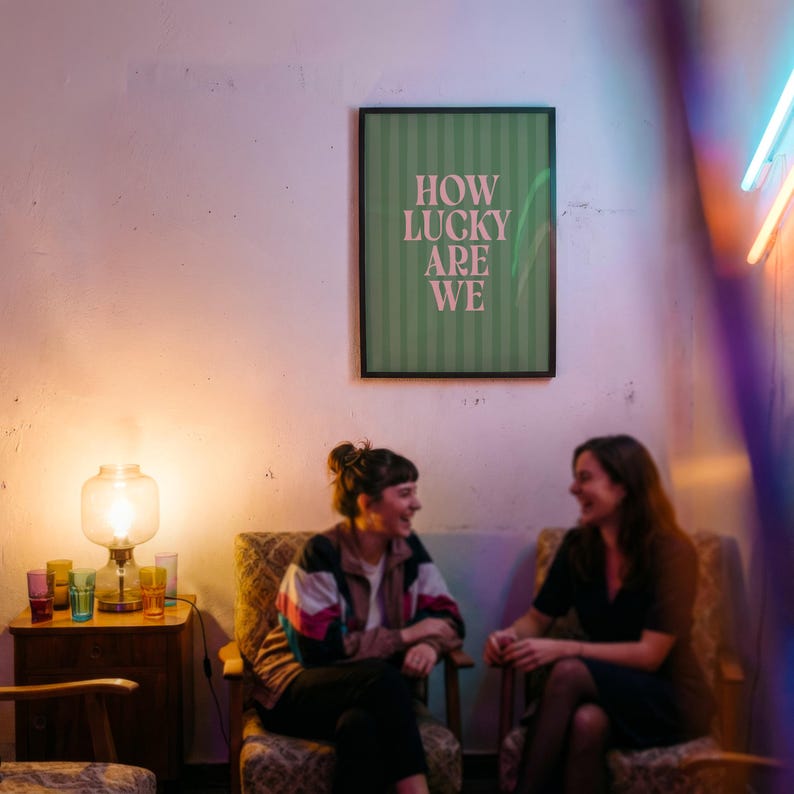 May include: Two people seated in patterned armchairs are laughing. A framed poster on the wall reads "HOW LUCKY ARE WE" in white text on a green striped background. A lit lamp and colorful glasses sit on a small wooden table.