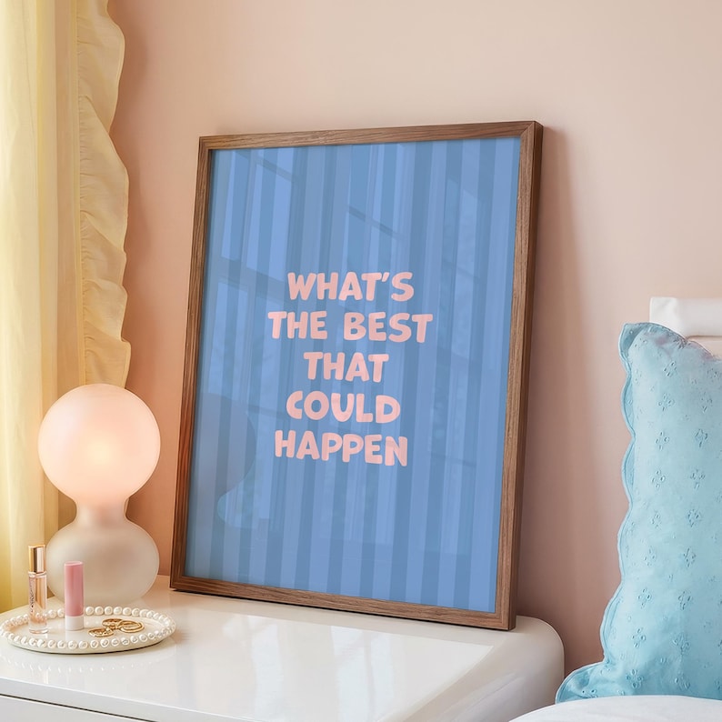 Puede incluir: Un estampado enmarcado con el texto "WHAT'S THE BEST THAT COULD HAPPEN" en letras rosas sobre un fondo azul a rayas. El marco es de madera. El estampado se exhibe en una mesita de noche blanca.