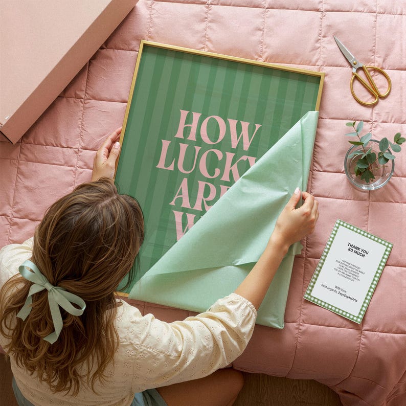 May include: A framed art print with the text "HOW LUCKY ARE WE" in pink letters on a green striped background. The frame is gold. The print is being unwrapped on a pink comforter. A small vase with greenery and a pair of gold scissors are nearby.