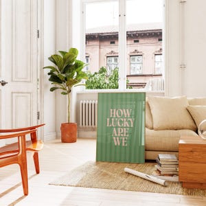 May include: A framed green poster with vertical stripes and the text "HOW LUCKY ARE WE" in pink. The poster is leaning against a beige sofa in a bright room with a large window and a potted plant.