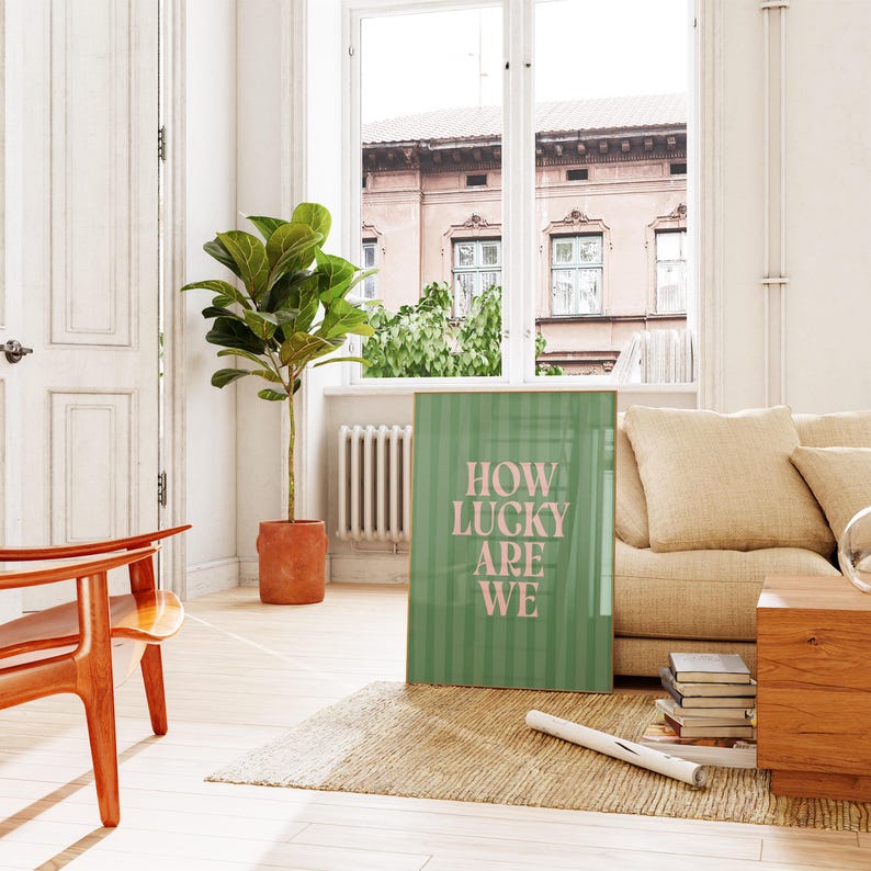 May include: A framed green poster with vertical stripes and the text "HOW LUCKY ARE WE" in pink. The poster is leaning against a beige sofa in a bright room with a large window and a potted plant.