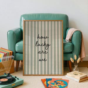 Puede incluir: Un cuadro enmarcado con rayas verticales verdes claras y blancas, con el texto "how lucky are we" en escritura negra. El cuadro se exhibe cerca de un sillón verde azulado, un tocadiscos y discos de vinilo.