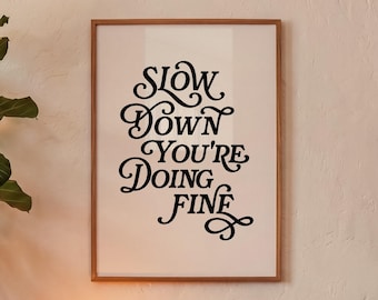 May include: A rectangular wall art print in a thin wooden frame featuring black decorative lettering on a cream background. The text reads: SLOW DOWN YOU'RE DOING FINE, written in a stylized, flowing script with ornate flourishes on the letters.