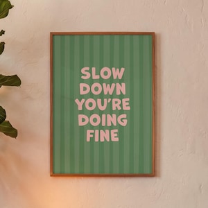 Puede incluir: Una lámina de arte mural enmarcada con un fondo verde y rayas verticales en un tono de verde más oscuro. El texto SLOW DOWN YOU'RE DOING FINE está impreso en el centro con una tipografía sans-serif rosa claro, negrita y redondeada.