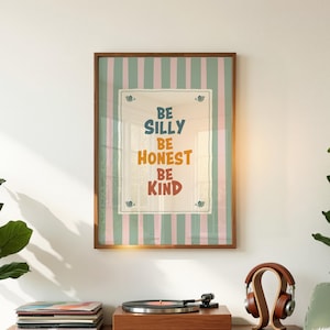 Puede incluir: Un cuadro enmarcado con las palabras "Be Silly, Be Honest, Be Kind" en azul, naranja y rojo. El fondo tiene rayas verticales rosas y verdes. El marco es marrón.