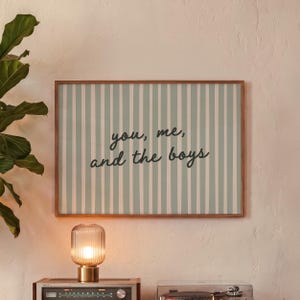 May include: A rectangular wall art print in a wooden frame featuring a light sage green and white vertical striped background with the black cursive text you, me, and the boys centered on the print. The design is clean and simple in its layout.