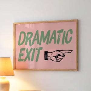 Puede incluir: Un estampado enmarcado con las palabras "DRAMATIC EXIT" en verde, con una mano negra que apunta hacia la derecha sobre un fondo rosa. El marco es de madera clara. El estampado está montado en una pared blanca.