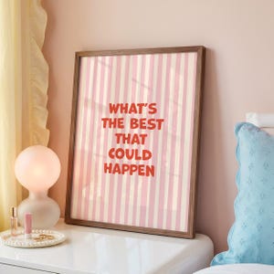 May include: A framed print with a pink and white striped background and the text "WHAT'S THE BEST THAT COULD HAPPEN" in red. The frame is a dark brown color. The print is displayed on a white nightstand.