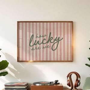 Puede incluir: Arte de pared enmarcado con un fondo a rayas rosas y blancas. La obra de arte presenta la frase "how lucky are we" en una fuente verde cursiva. El marco es de color marrón cálido.
