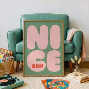 Op de afbeelding: Een rechthoekige wandkunstprint in een houten lijst met een saliegroene achtergrond en grote, afgeronde, lichtroze bubbelletters die het woord NICE spellen, met het woord BUM in kleinere, vetgedrukte, oranjerode letters eronder.