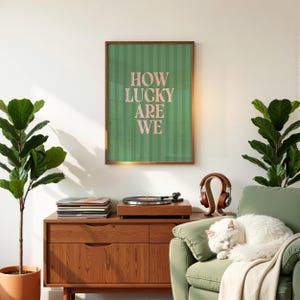 May include: A framed print with the text "HOW LUCKY ARE WE" in pink letters on a green striped background. The print hangs above a wooden record player and a stack of vinyl records. A white cat rests on a green armchair.