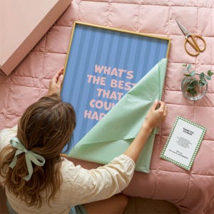 Puede incluir: Una impresi&oacute;n art&iacute;stica enmarcada con el texto "WHAT'S THE BEST THAT COULD HAPPEN" en letras rosas sobre un fondo a rayas azules. El marco es dorado. Una persona est&aacute; desenvolviendo la impresi&oacute;n sobre un edred&oacute;n rosa. Tambi&eacute;n se ven una tarjeta peque&ntilde;a y unas tijeras.