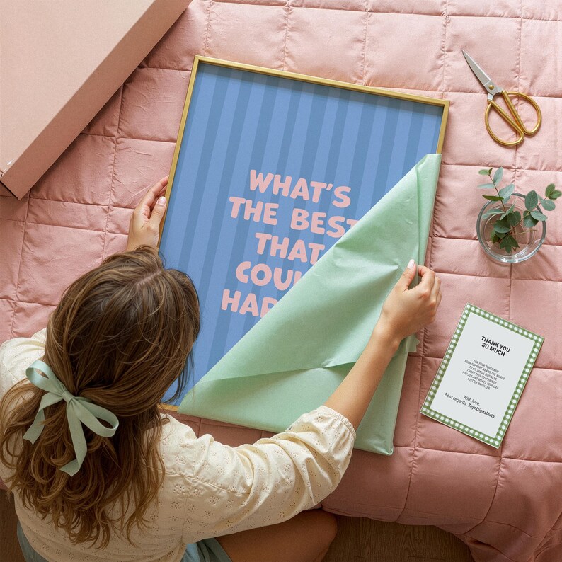 Puede incluir: Una impresi&oacute;n art&iacute;stica enmarcada con el texto "WHAT'S THE BEST THAT COULD HAPPEN" en letras rosas sobre un fondo a rayas azules. El marco es dorado. Una persona est&aacute; desenvolviendo la impresi&oacute;n sobre un edred&oacute;n rosa. Tambi&eacute;n se ven una tarjeta peque&ntilde;a y unas tijeras.