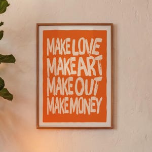 以下が含まれることがあります： 鮮やかなオレンジ色の背景に、太字の手描き風フォントで「MAKE LOVE MAKE ART MAKE OUT MAKE MONEY」という白いテキストが書かれた、長方形の額入りウォールアートプリント。プリントは薄い明るい色の木製フレームに収められています。