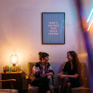 Puede incluir: Dos personas sentadas en sillones estampados conversan. Un p&oacute;ster enmarcado en la pared dice "WHAT'S THE BEST THAT COULD HAPPEN" en texto rosa. Una l&aacute;mpara y vasos coloridos est&aacute;n en una mesa auxiliar. La escena est&aacute; iluminada con luz c&aacute;lida.