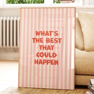 May include: A framed print with a pink and cream striped background. Bold red text reads "WHAT'S THE BEST THAT COULD HAPPEN". The artwork is displayed in a room with a window, radiator, and a beige sofa.