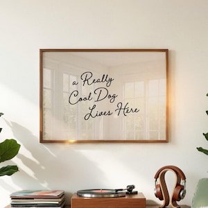 以下が含まれることがあります： 「a Really Cool Dog Lives Here」という筆記体のテキストが入った額入りのプリント。茶色のフレームは、レコードプレーヤーとヘッドフォンの上の白い壁に表示されています。