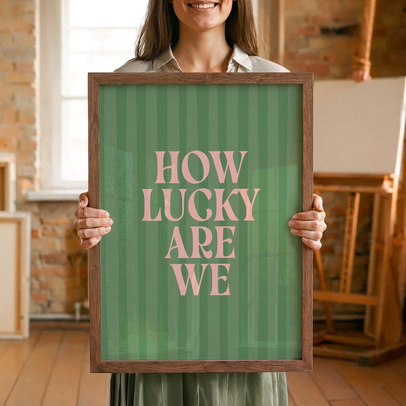 May include: A framed art print with a green striped background and the text "HOW LUCKY ARE WE" in pink. The frame is a dark brown color. The art print is held by a person.