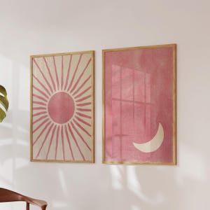 Può includere: Due stampe artistiche incorniciate. Una presenta un motivo a raggiera rosa su sfondo crema. L'altra ha uno sfondo rosa con una luna crescente bianca. Entrambe sono in cornici di legno marrone chiaro.