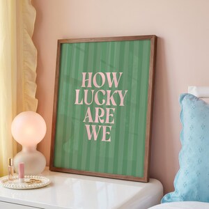 May include: A framed art print with the text "HOW LUCKY ARE WE" in pink letters on a green striped background. The frame is made of wood. The print is displayed on a white surface.