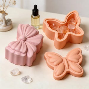 Może przedstawiać: Kolekcja dekoracyjnych pojemników w kształcie kokardy i motyli. Jeden pojemnik jest wypełniony kryształami. Pojemniki są w odcieniach różu i brzoskwini. Obecna jest również mała szklana butelka z zakraplaczem.