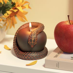 Può includere: Una candela decorativa a forma di mela, con un serpente marrone e dettagli arancioni. Uno stoppino acceso è al centro. Una mela rossa e girasoli sono sullo sfondo. Un oggetto di arredamento unico.