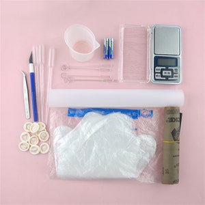 May include: A set of tools for crafting and making resin jewelry. The set includes a small digital scale, a measuring cup, a pair of tweezers, a pair of scissors, a set of droppers, a set of stirring sticks, a roll of plastic wrap, a pair of disposable gloves, and a roll of sandpaper.