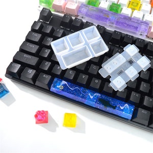 May include: A black keyboard with a blue and white resin keycap, a clear silicone mold for making resin keycaps, and a clear silicone mold for making resin keycap sets. There are also several small resin keycaps in different colors.