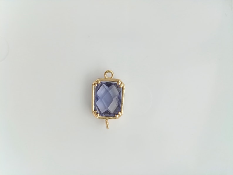 Puede incluir: Un colgante dorado con una piedra preciosa de amatista de forma cuadrada. El colgante tiene un bucle en la parte superior para sujetarlo a una cadena.