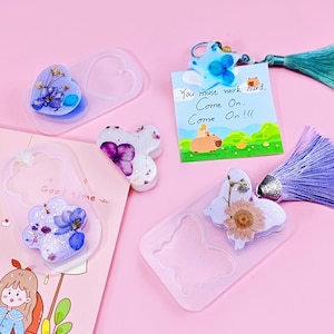 May include: A collection of resin crafts, including heart and butterfly-shaped pieces with dried flowers and glitter. A small card with the text "You must work hard, Come On. Come On!!!" is also present, along with a tassel.