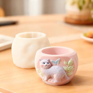 Può includere: Due piccoli vasi rotondi. Uno è di colore bianco sporco pieno. L'altro è rosa con un gatto bianco, una farfalla blu e un motivo di foglie verdi. Il gatto ha gli occhi blu ed è sdraiato.